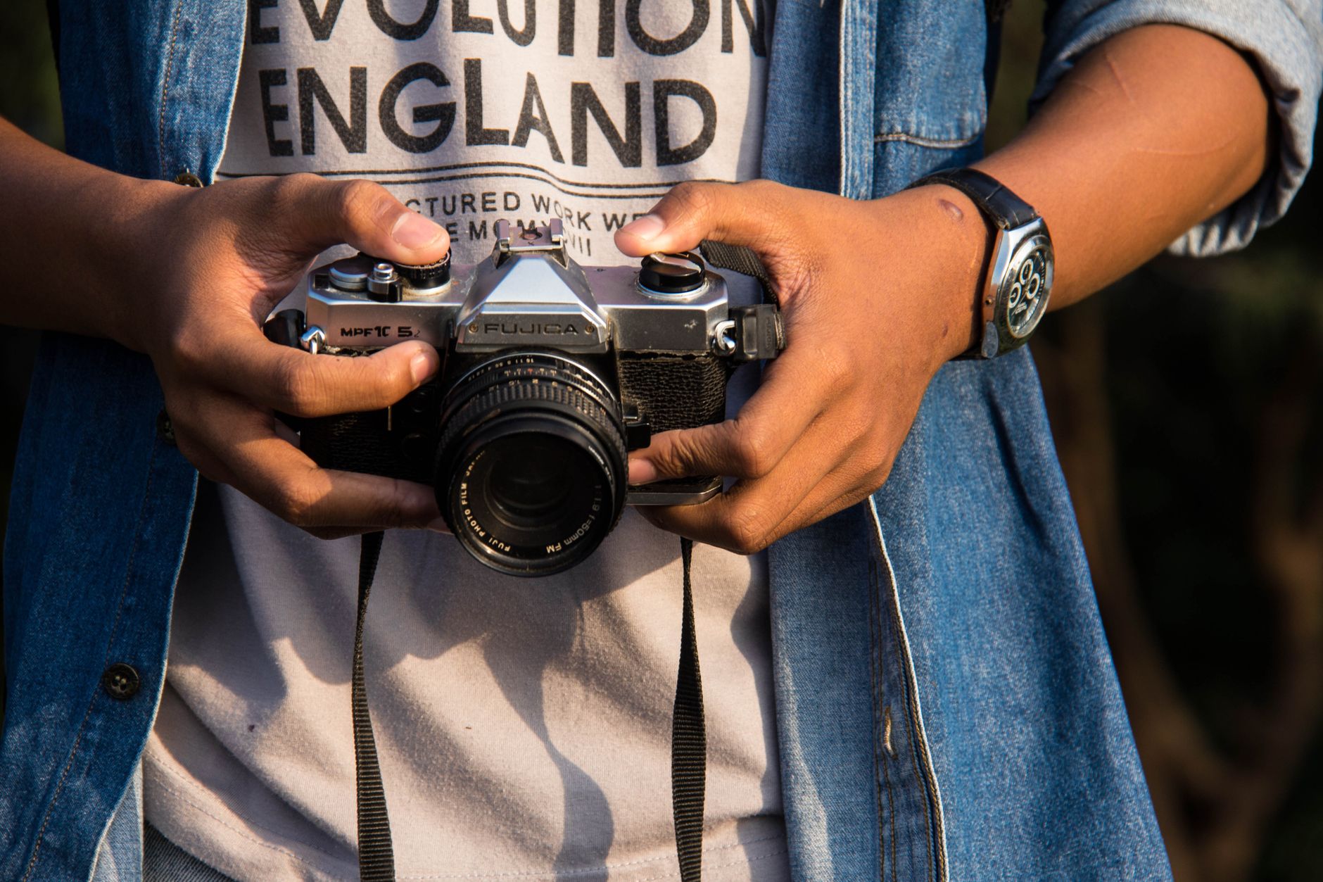 A person holding a vintage analog SLR camera outdoors, wearing a casual outfit.