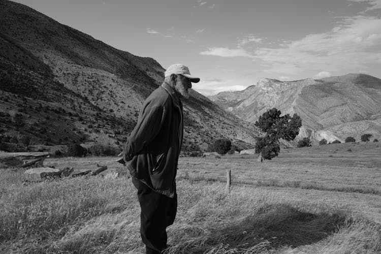 Grayscale Photo Of An Elderly Man With A Cap Standing On A Field