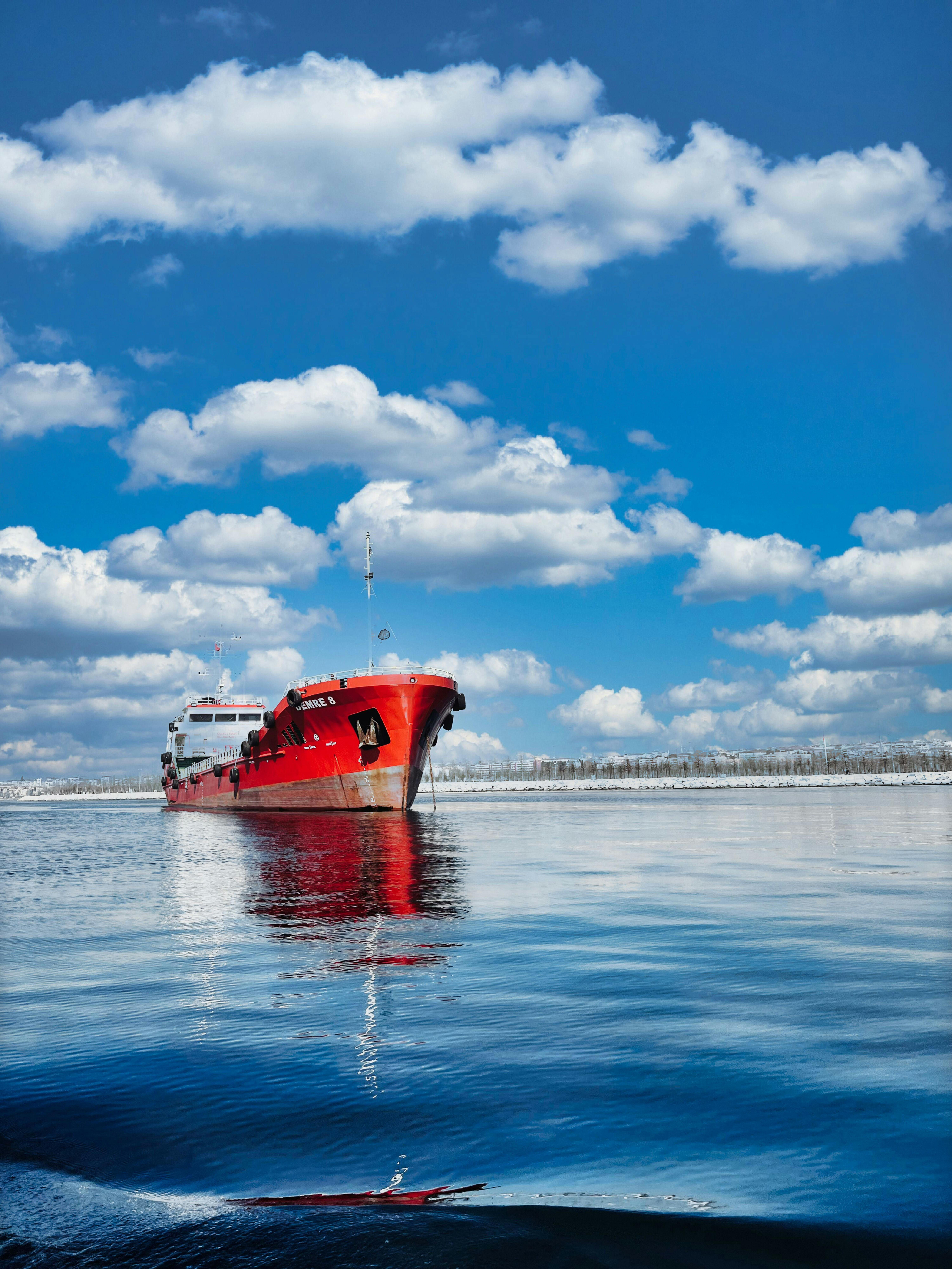 Free A striking red ship sails under blue skies with white clouds reflected in serene waters. Stock Photo