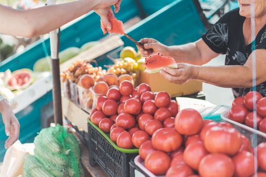 A vendor hands a watermelon slice at a vibrant outdoor market with various fruits.