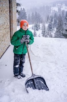 Young child in winter gear shoveling snow near a house with snowy forest backdrop.