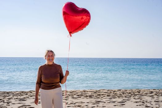 Smiling woman holding a heart-shaped balloon on a sunny beach.