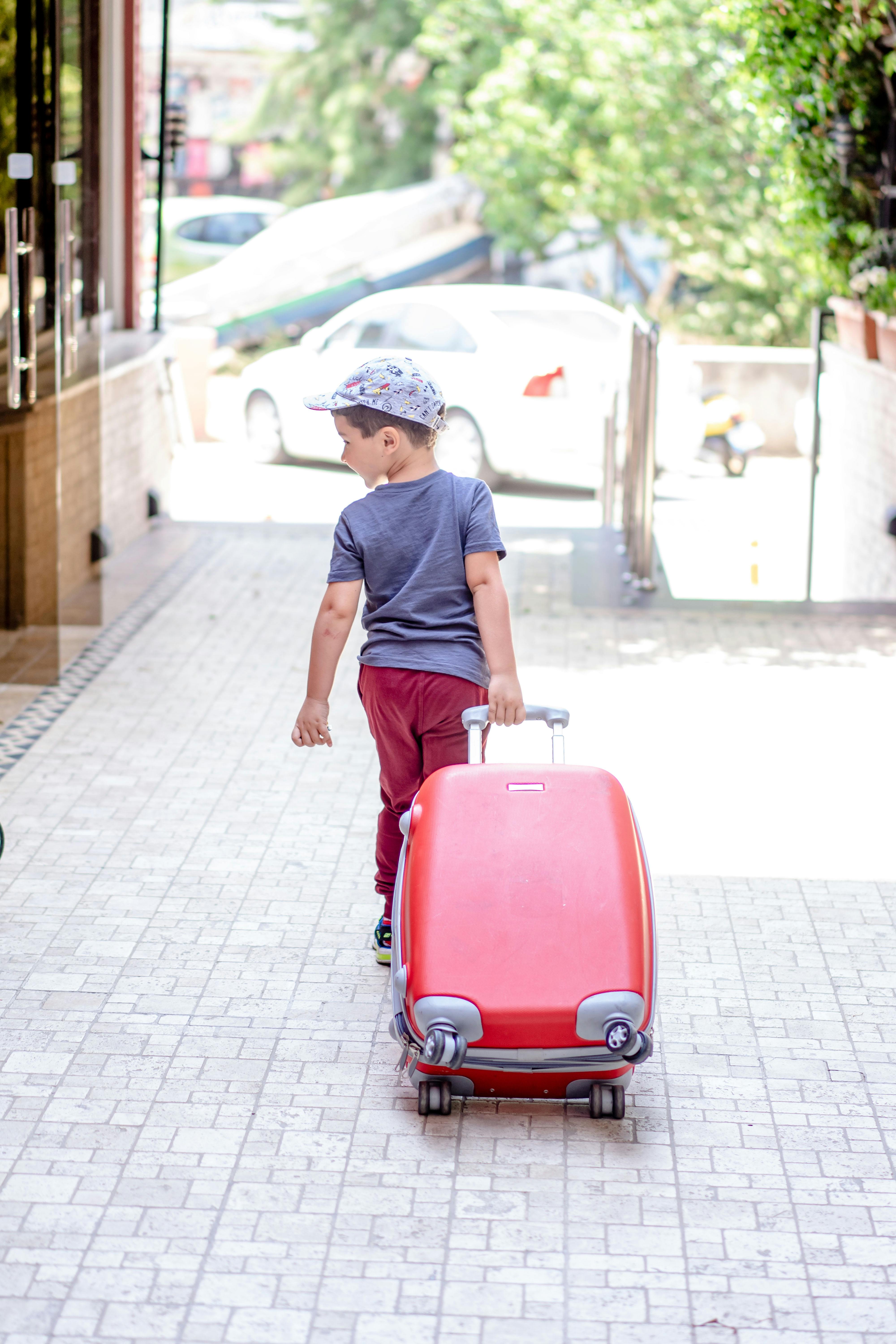 A young boy in a gray shirt and red pants pulling a suitcase outdoors.