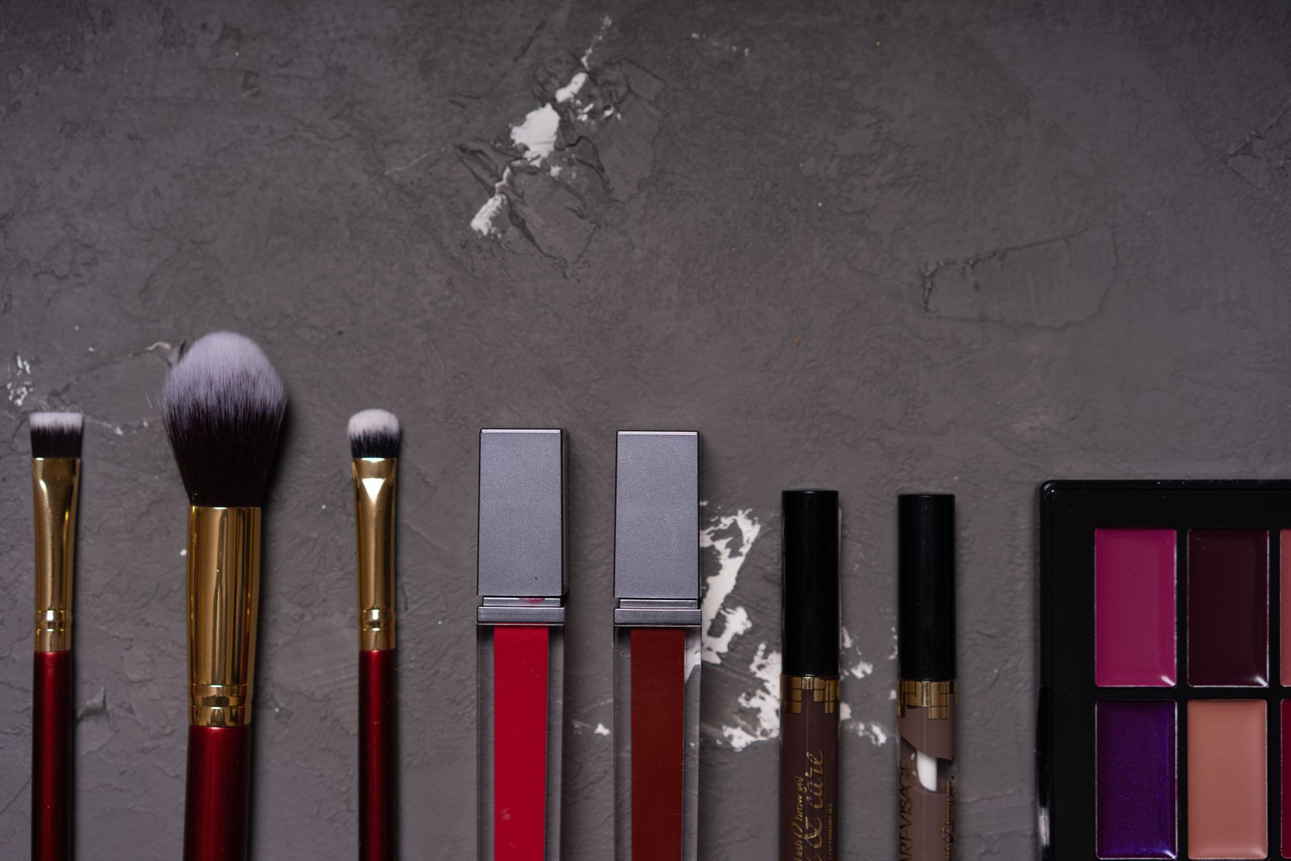 close-up flat lay of bridal makeup products and brushes including warm-toned eye shadow palette, nude and rose lip colors, and cream blush, arranged on ivory linen