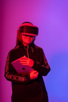 A teenage girl wearing a VR headset holds a tablet, enjoying a virtual experience in pink and blue lighting.