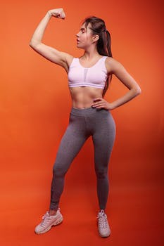 A fit woman in activewear confidently flexing on a vibrant orange background, symbolizing strength and wellness.
