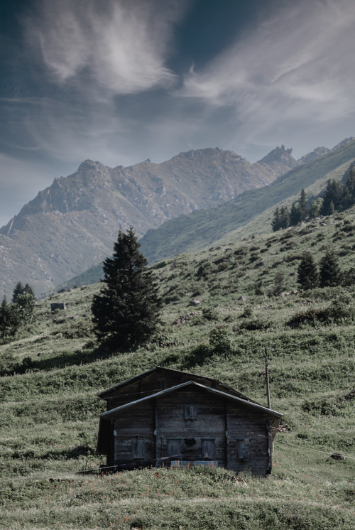 Free Charming wooden cabin nestled in a lush grassland with mountain backdrop and cloudy sky. Stock Photo