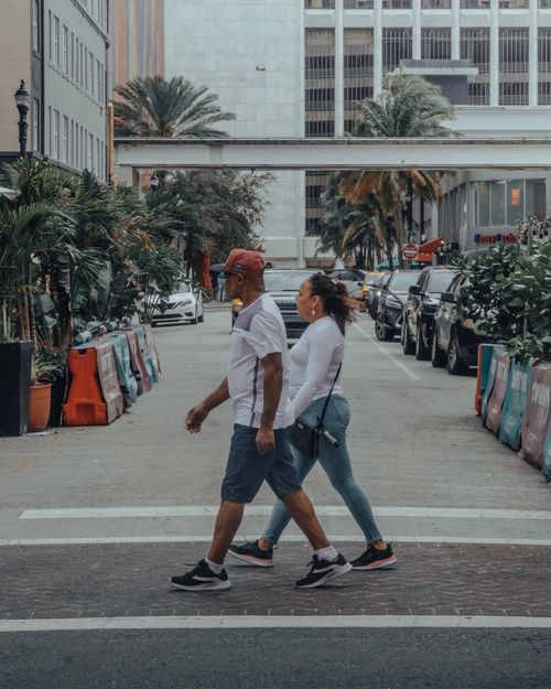 Free A couple walks in an urban setting surrounded by palm trees, buildings, and parked cars. Stock Photo