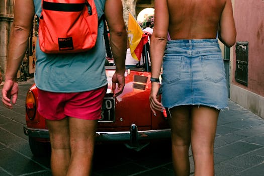 Back view of a man and woman walking down a street near a red vintage car, enjoying a sunny day.