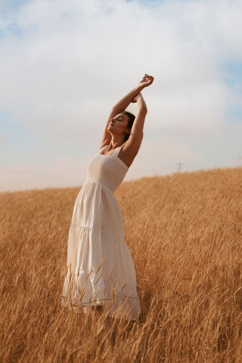 Free A woman in a white dress enjoying a peaceful moment in a golden wheat field under a bright sky. Stock Photo