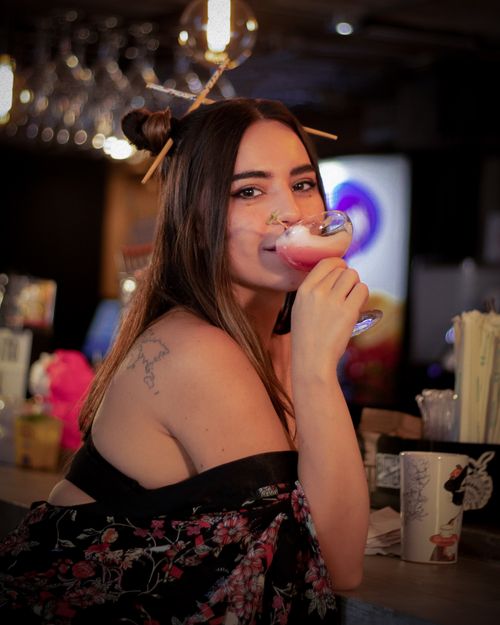Free Young woman sipping a cocktail with a smile, exuding style and elegance in a dimly lit bar. Stock Photo