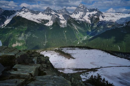 A breathtaking aerial photo of the snow-covered Rocky Mountains amidst lush greenery and clear blue skies.