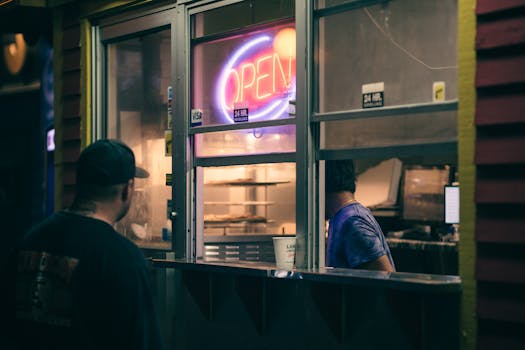 Man ordering food at a street stall with neon 'Open' sign at night.