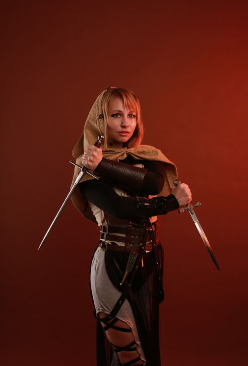 Free A woman in creative cosplay attire, holding swords in a dramatic studio setting. Stock Photo