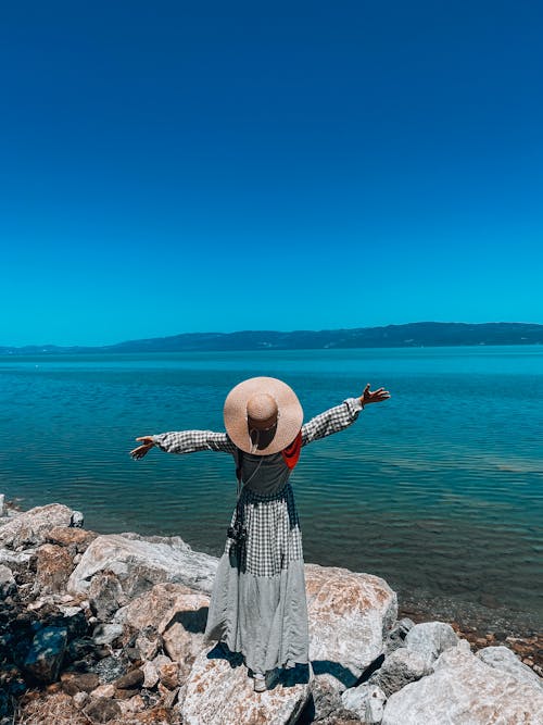 Back View of a Woman in Checkered Dress Wearing Sun Hat while Standing on Rocks in Front of the Blue Ocean