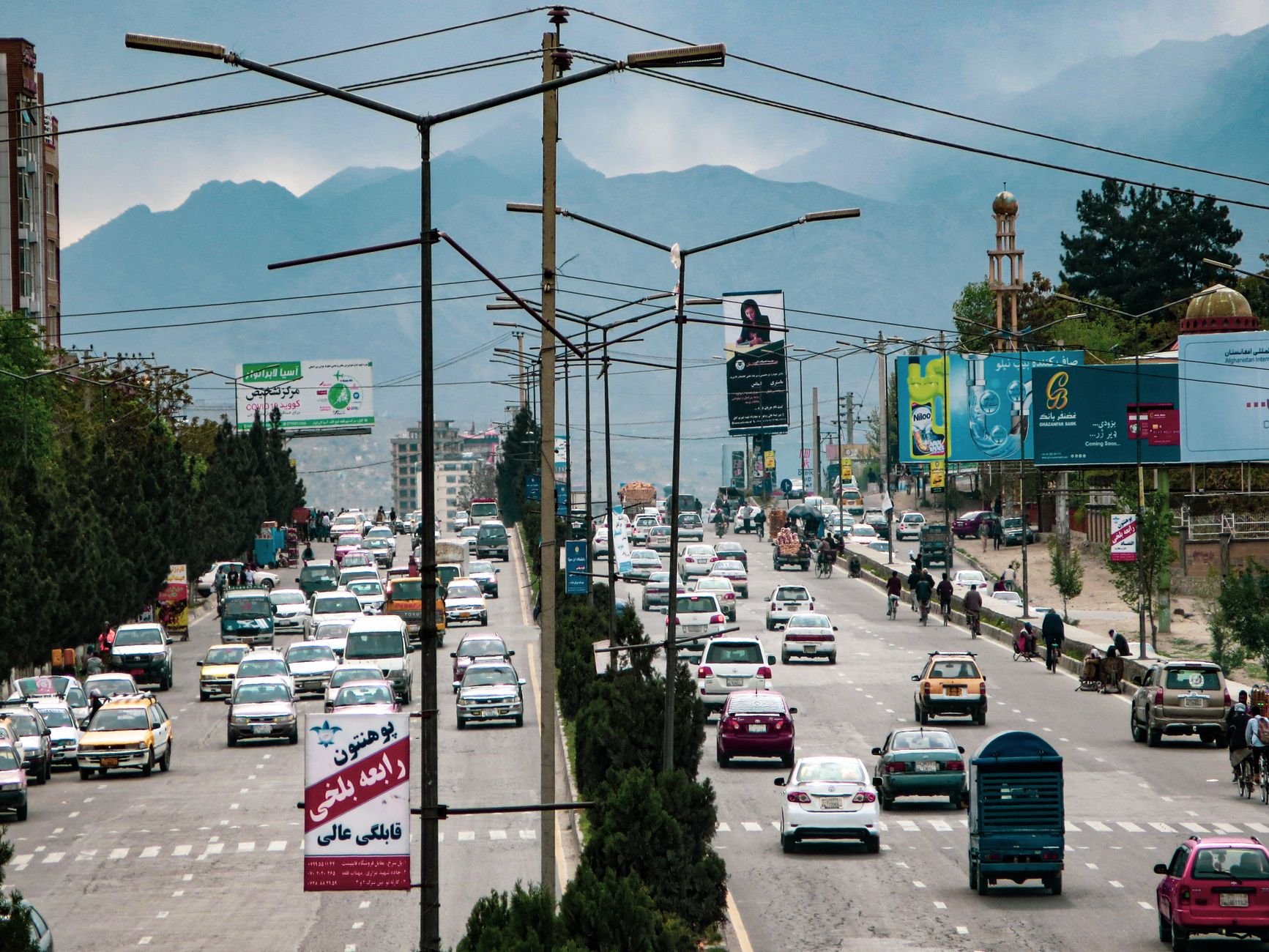 Street view of traffic and billboards in Kabul, Afghanistan, with a mountainous backdrop.
