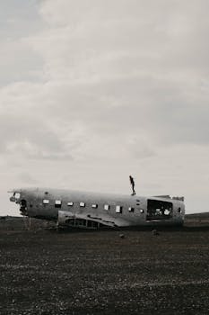 A lone person stands on a wrecked aircraft fuselage in an empty, moody landscape.