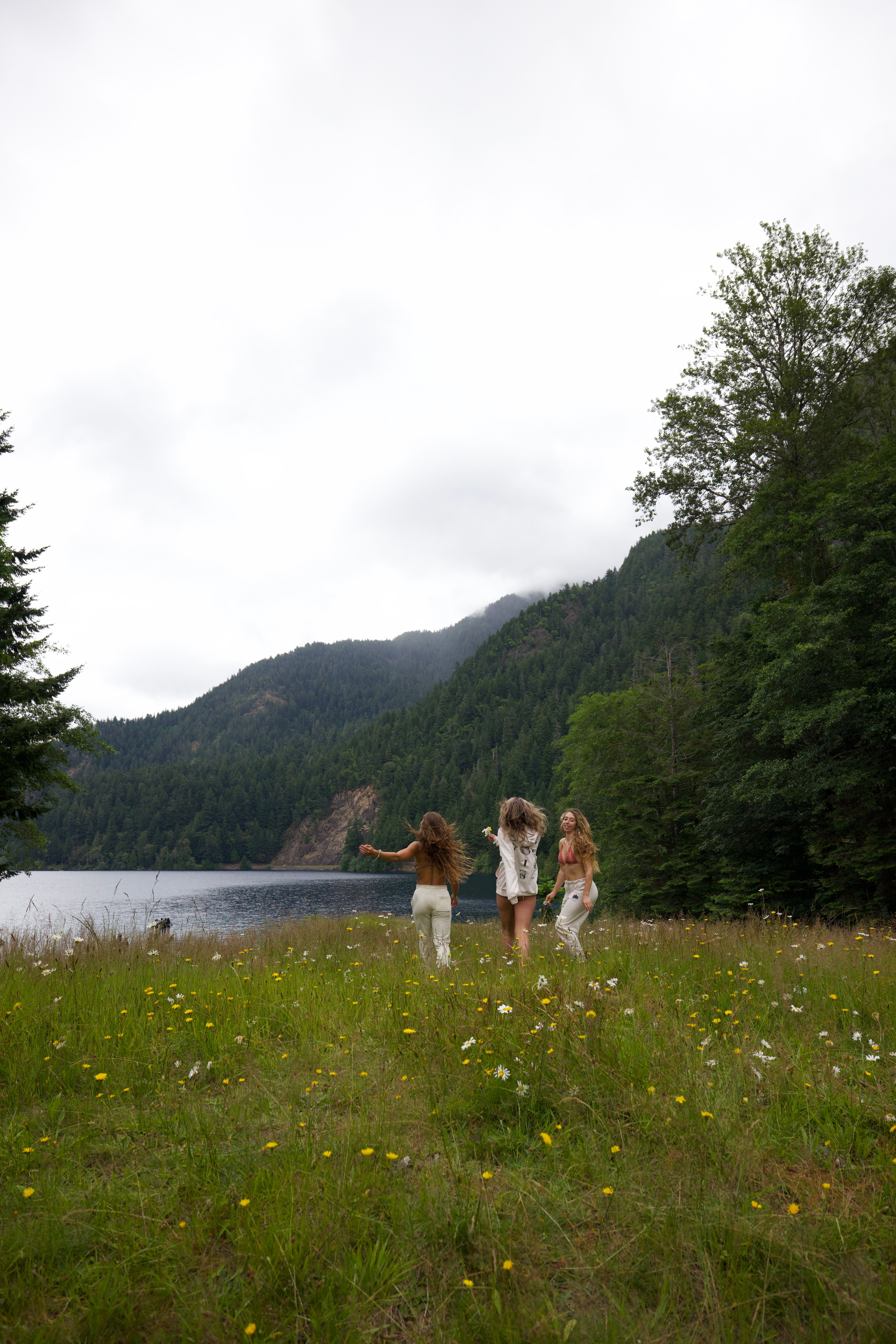 Three women running in a wildflower meadow by a mountain lake, capturing nature's beauty.