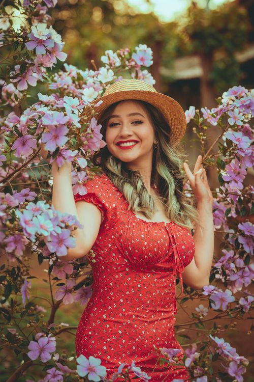 Free Joyful woman in a red dress and hat smiling amidst purple flowers outdoors. Stock Photo