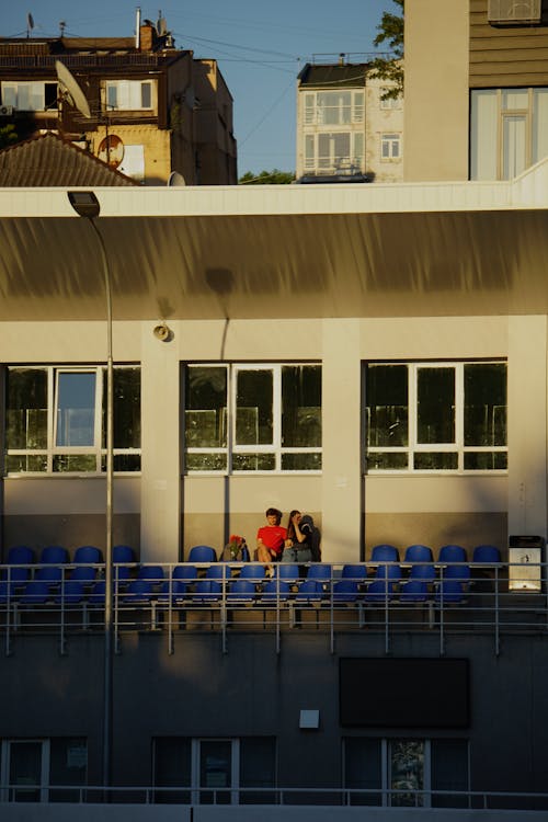 A Couple Sitting on Blue Plastic Seats Outside of a Building in City 