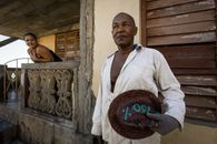A man in a white shirt and glove holds a hat outside a rustic house as a woman observes from the balcony.
