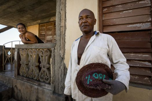 A man in a white shirt and glove holds a hat outside a rustic house as a woman observes from the balcony.