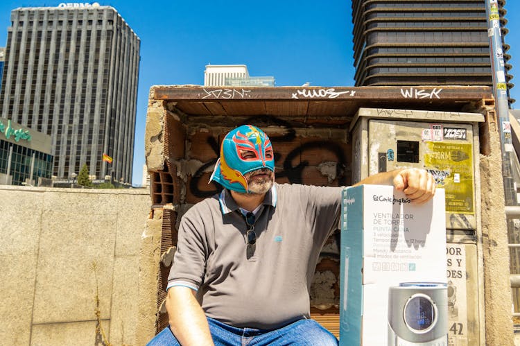 Man Wearing Colorful Luchador Mask Sitting Next To Ventilator Box