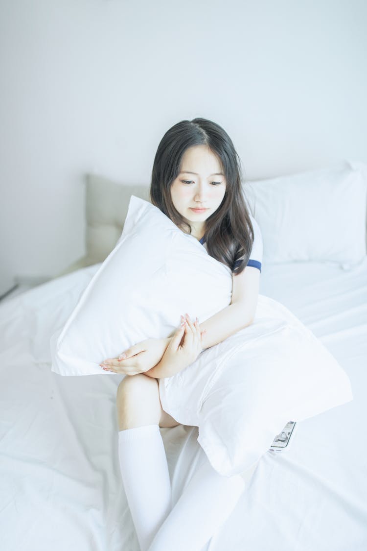 Girl Sitting On White Bed While Holding Her White Pillow