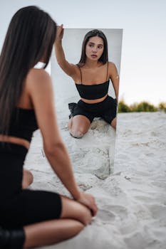 Woman with long hair posing in front of a mirror on a sandy beach during the day.