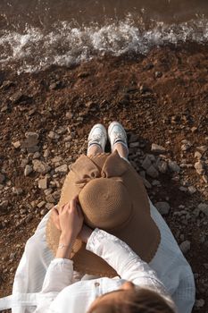 A woman in a white dress relaxes on a rocky shoreline wearing a brown hat, enjoying the coastal scenery.
