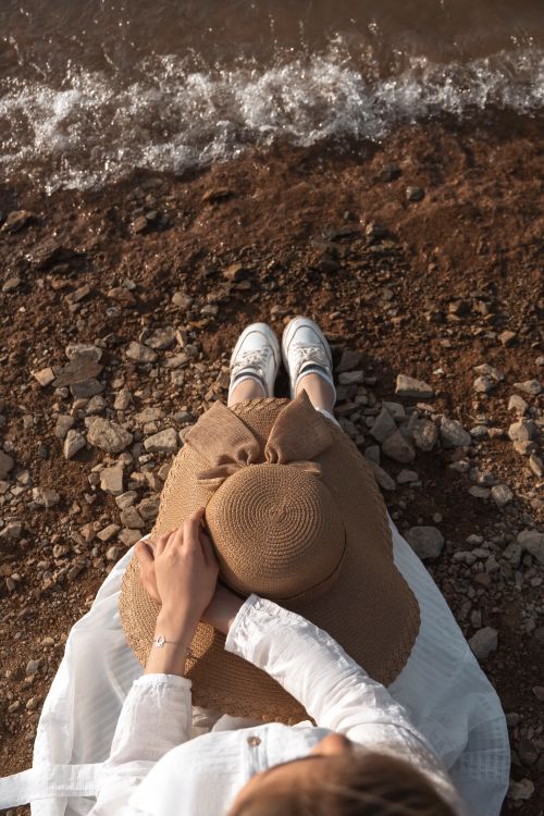 Free A woman in a white dress relaxes on a rocky shoreline wearing a brown hat, enjoying the coastal scenery. Stock Photo