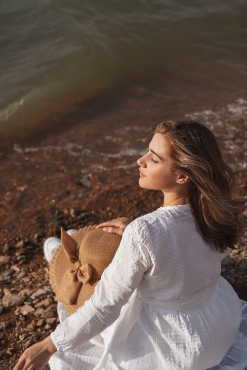 Free A woman in white enjoys the sun at a quiet seaside shore. Stock Photo