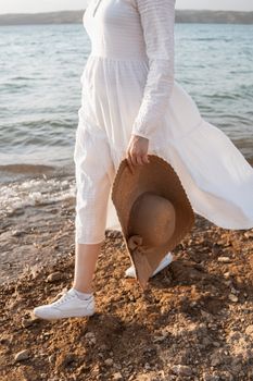 Elegant woman in a white dress holding a sun hat, walking on the beach shore on a sunny day.