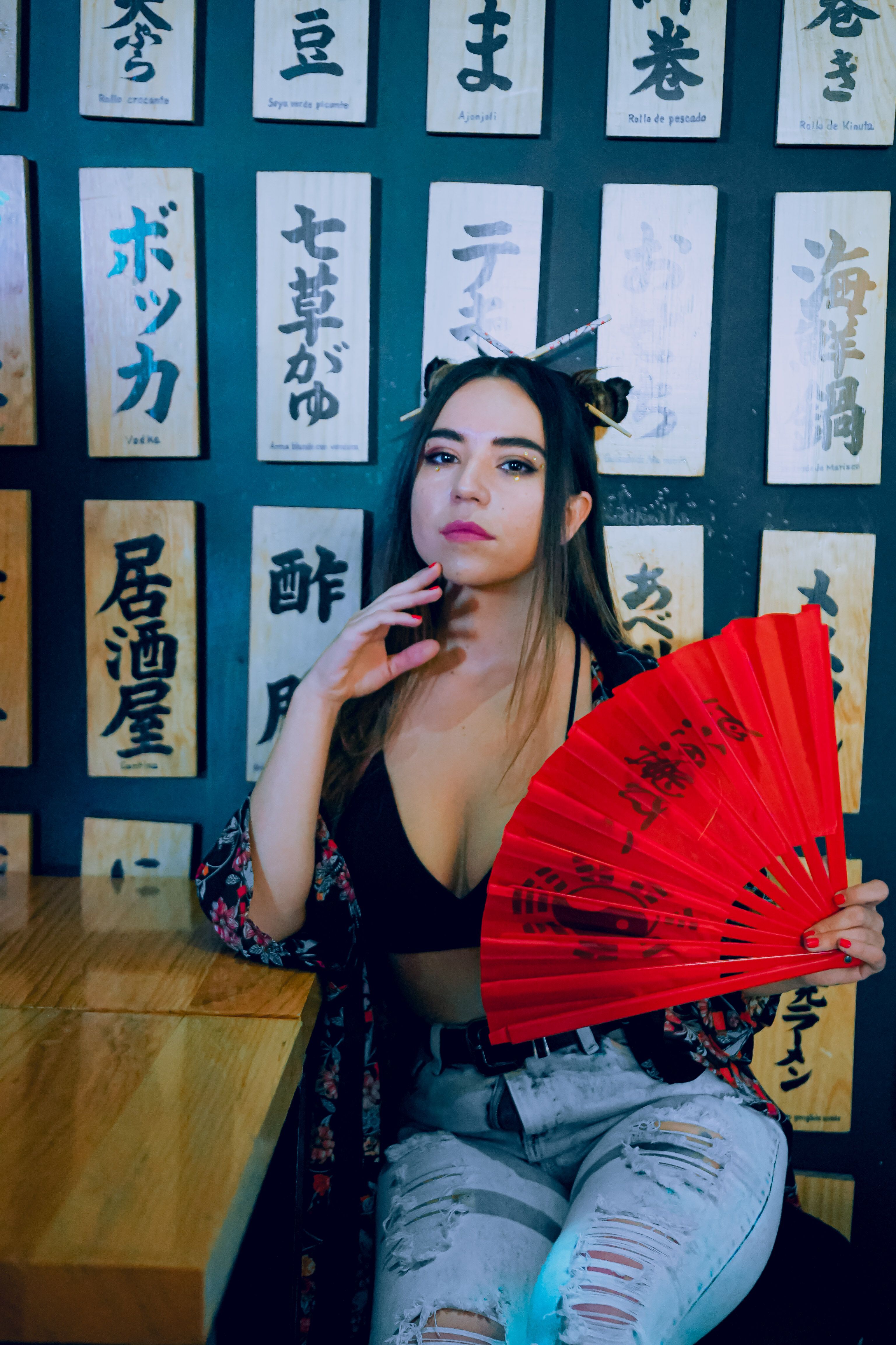A stylish woman in ripped jeans and a printed shirt holds a red fan in a modern cafe with Japanese decor.