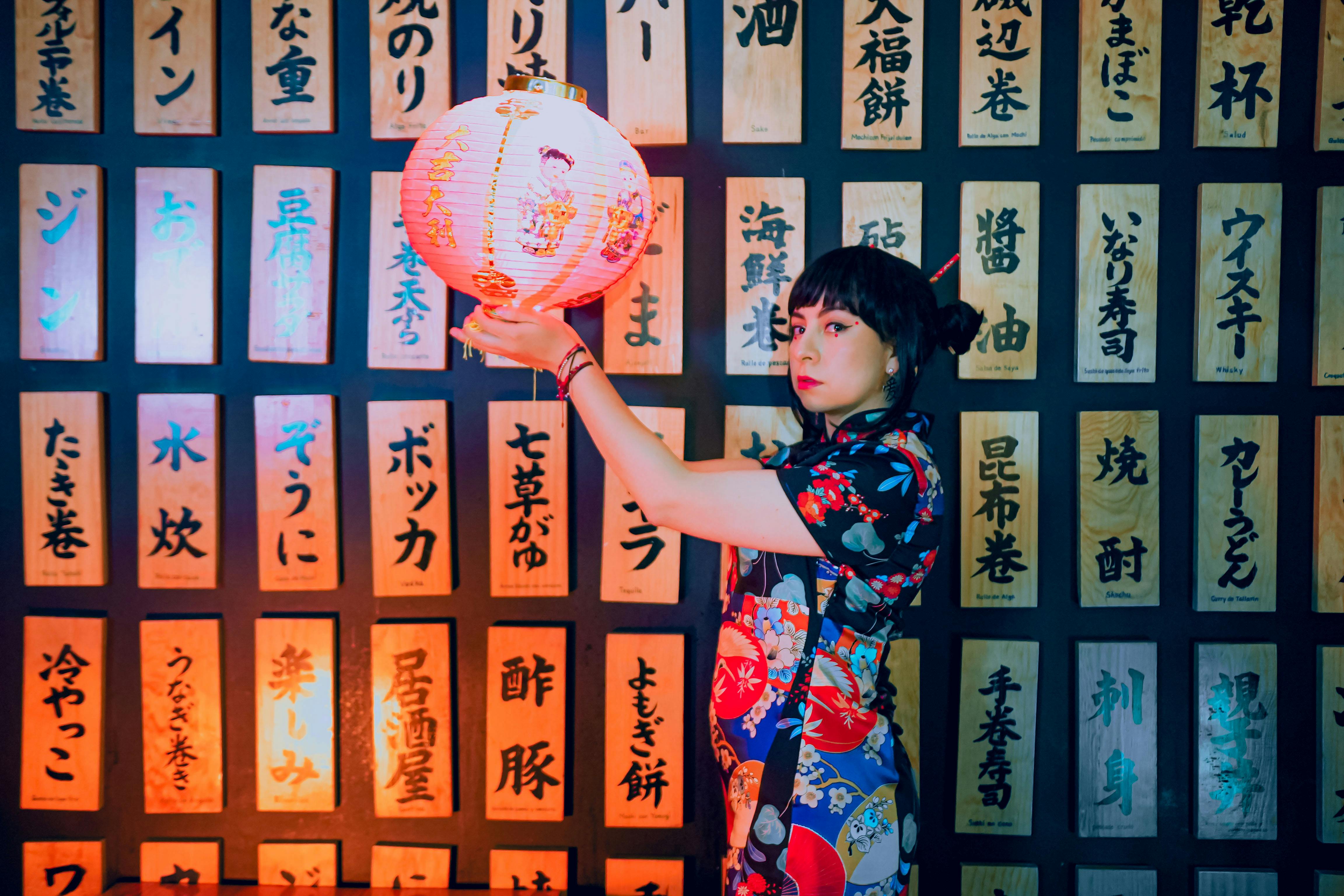 A woman in traditional attire holding a Japanese lantern against a backdrop of Japanese writings.