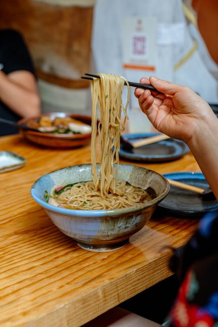 A tangle of pale yellow ramen noodles being lifted with chopsticks over a bowl of broth, showing their glossy, springy strands.