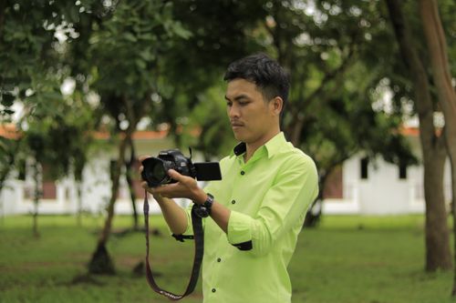 Free Asian male photographer in green shirt using camera outdoors in lush setting. Stock Photo