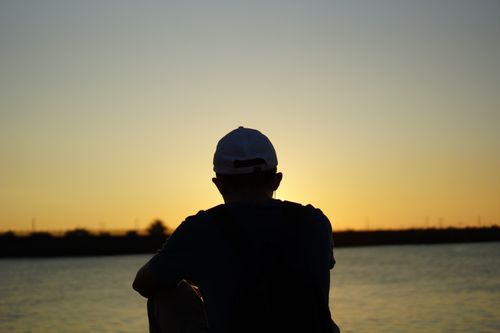 Free A man in silhouette wearing a cap, sitting by Hualien River at sunset in Hualien City, Taiwan. Stock Photo