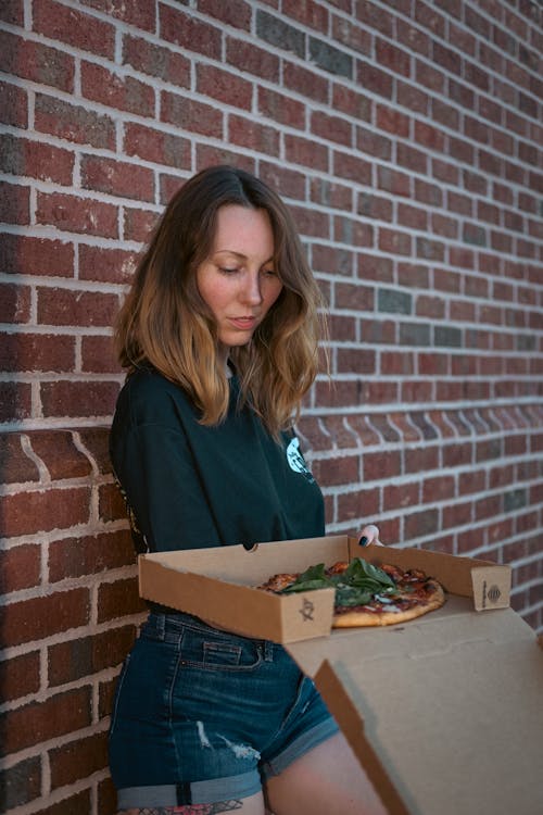 Woman Leaning on Brick Wall Holding a Box of Pizza