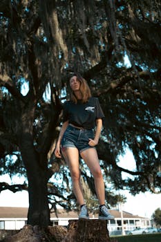 Woman in denim shorts on a tree stump, under a large tree during daytime.