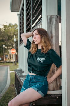 A relaxed portrait of a woman leaning against a wooden structure outdoors in casual denim shorts.