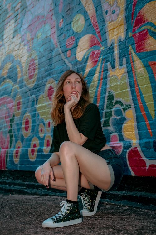 Free A thoughtful woman crouching near a colorful mural on a brick wall, wearing casual sneakers and shorts. Stock Photo