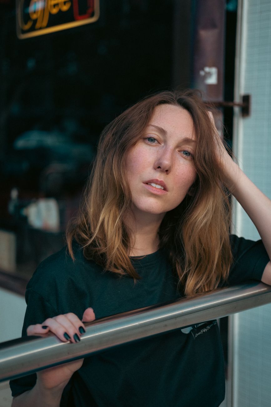 A woman with long hair leans on a railing outside a cafe, exuding a relaxed vibe.