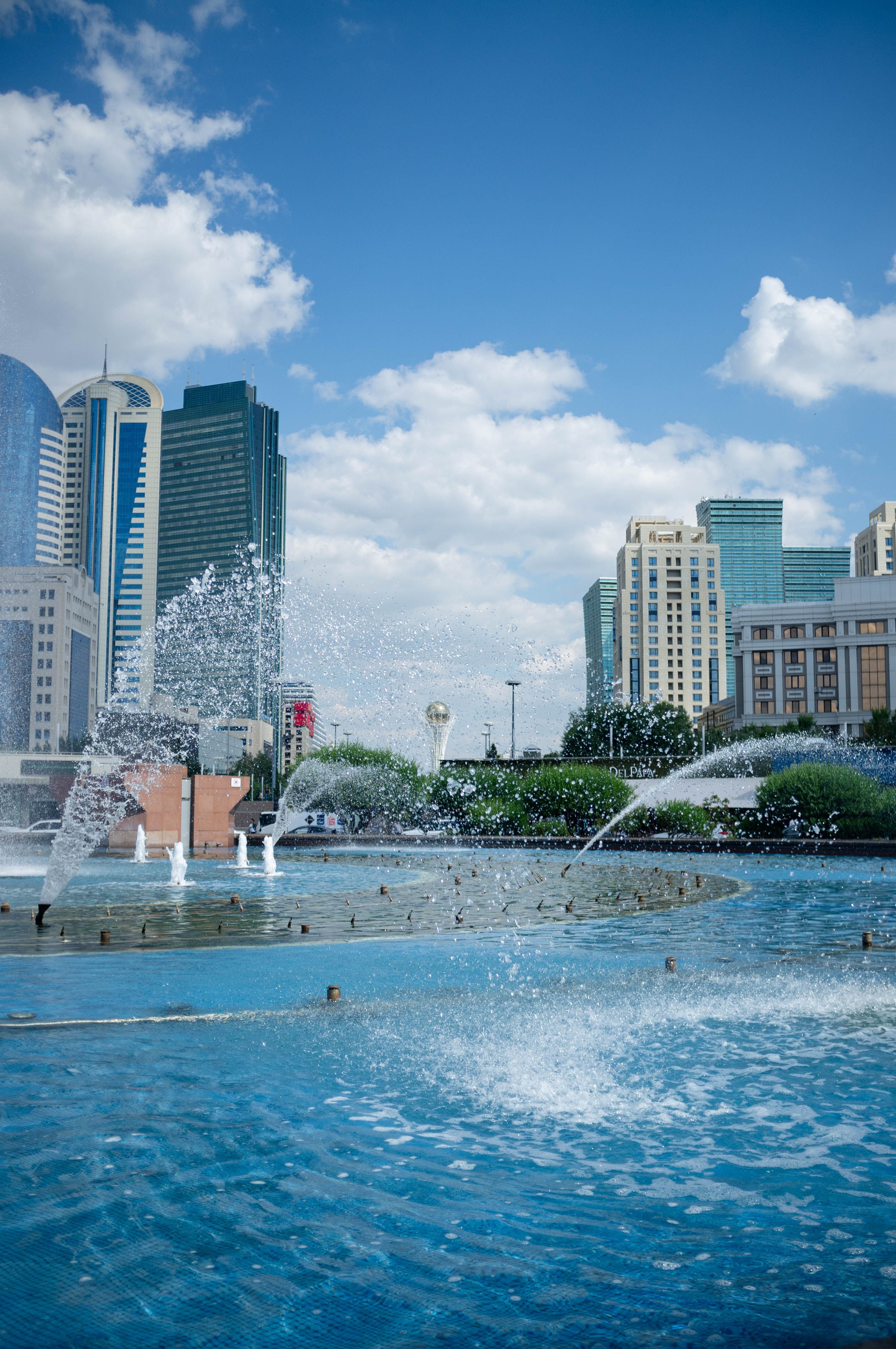Vibrant cityscape of Nur-Sultan with modern skyscrapers and a dynamic fountain under a blue sky.