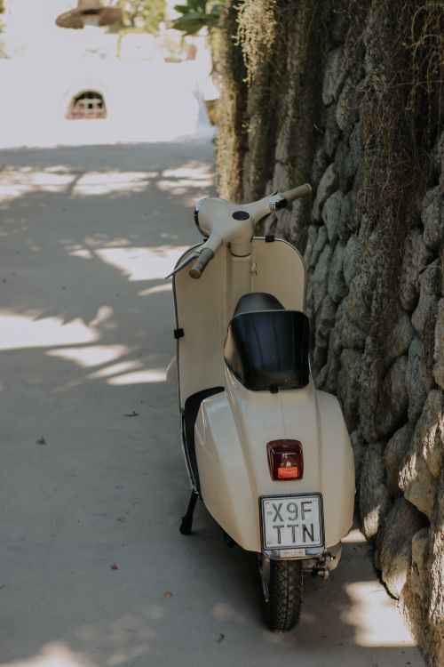 Free A vintage beige scooter parked on a sunlit cobblestone alley, next to a stone wall. Stock Photo