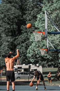 Shirtless player shooting a basketball on an outdoor court amid trees.