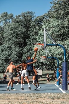 Men playing basketball outdoors under a blue sky with green trees in the background.