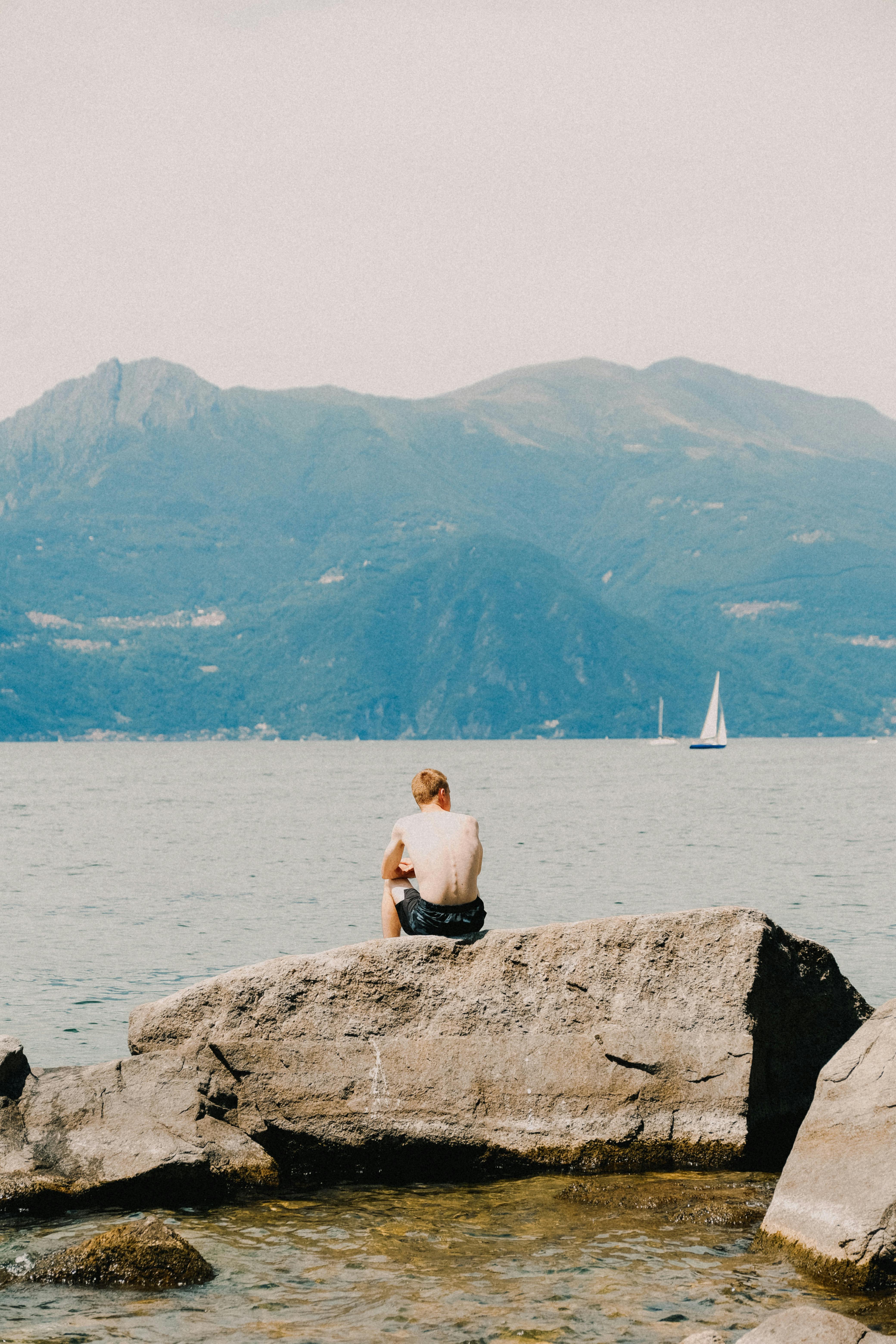 A man sits on rocks by the sea, overlooking mountains and a distant sailboat.