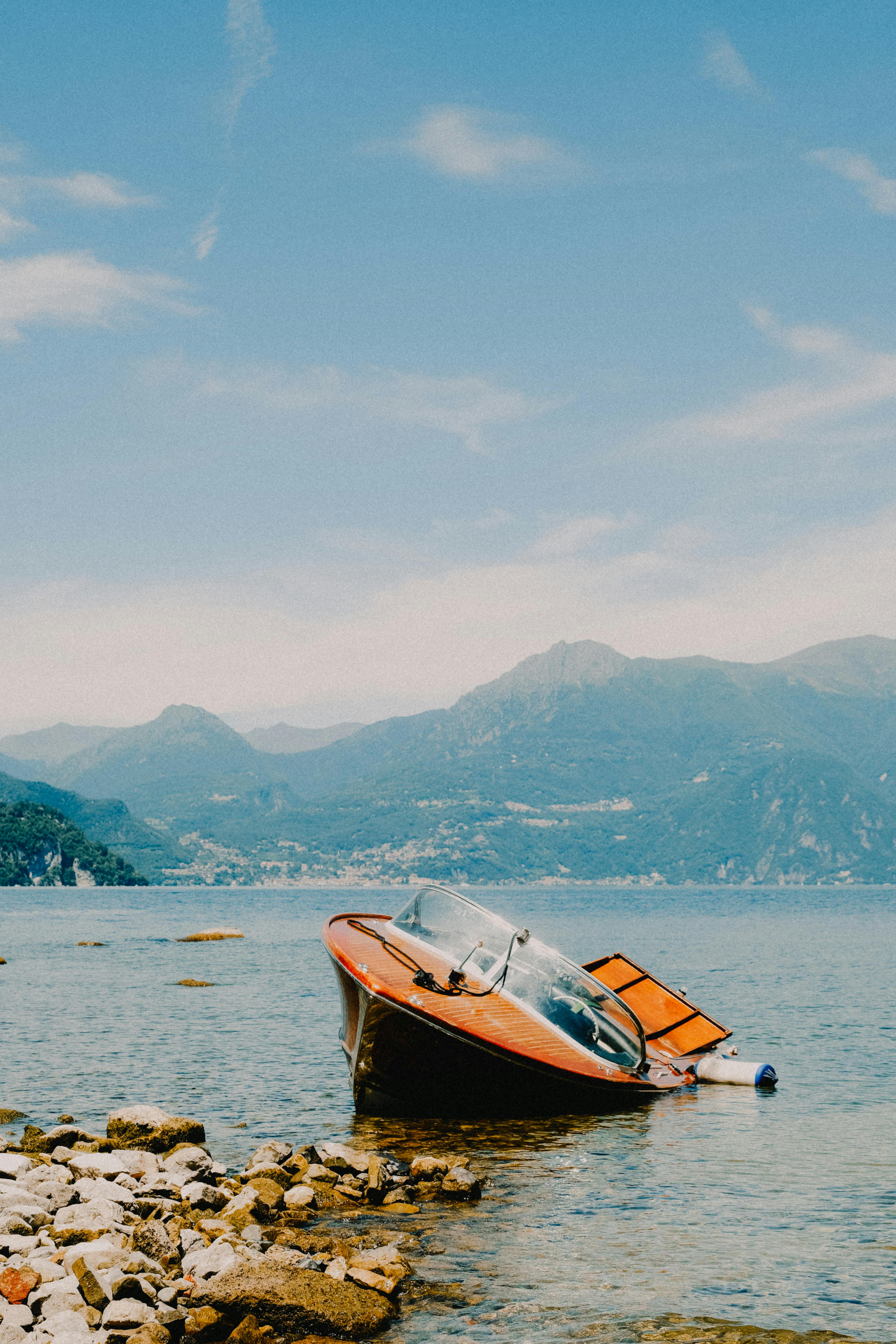 An abandoned motorboat rests on a rocky shore with mountains in the background, under a clear sky.