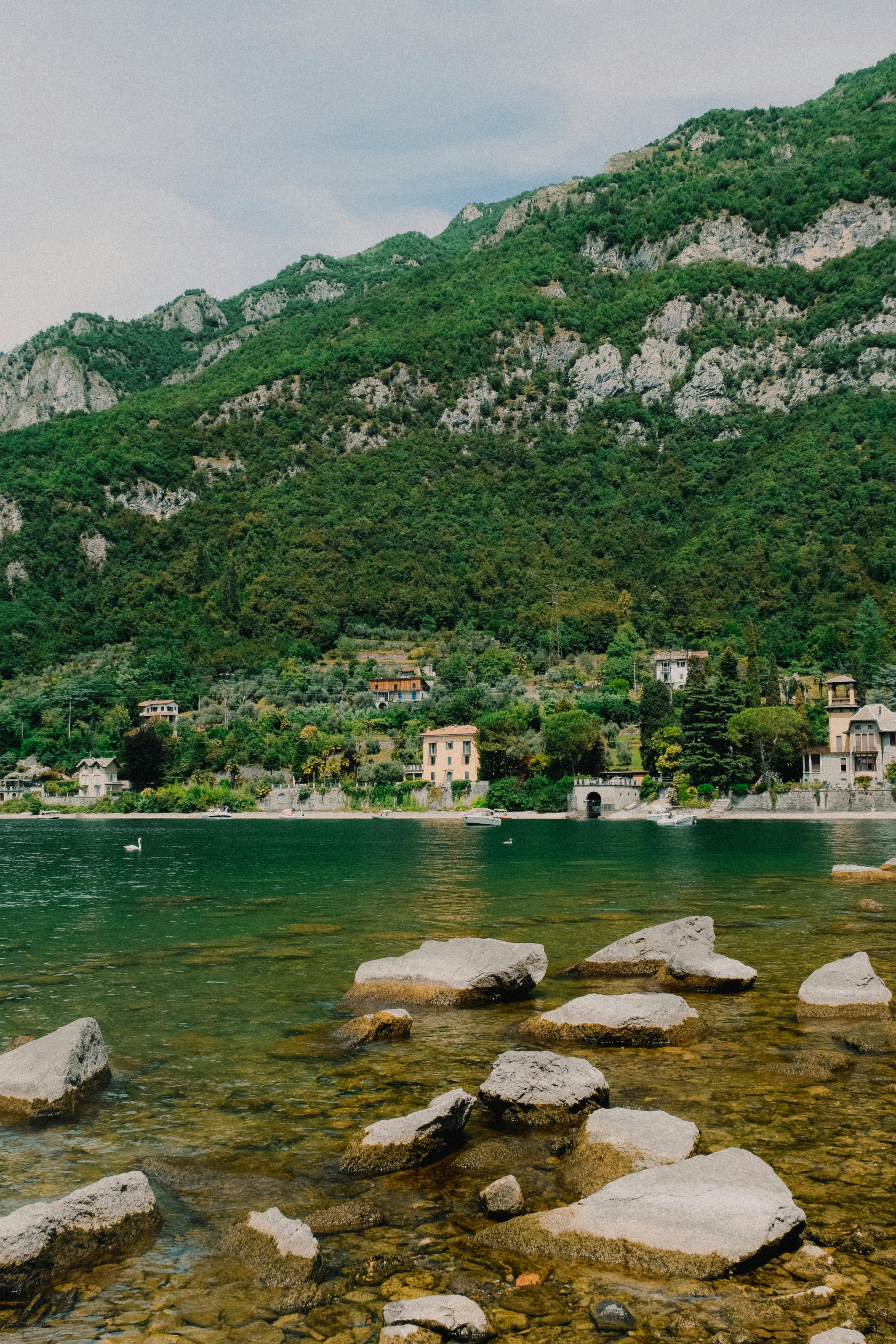 Tranquil scene of a green mountain, clear lake, and rocky shore under a cloudy sky.